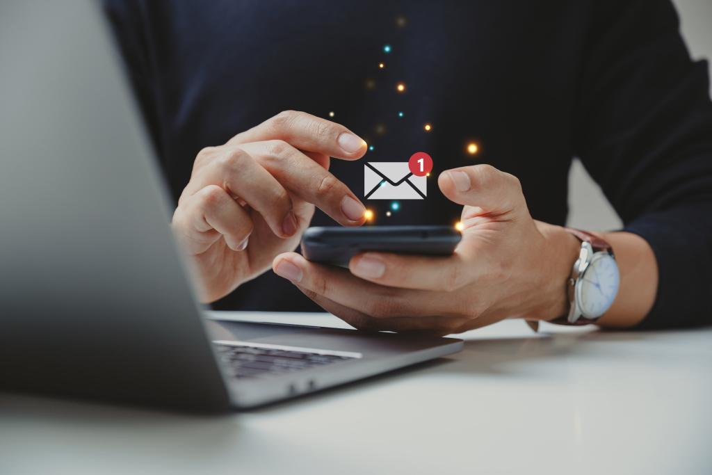 closeup of a person with a laptop holding a phone with an email notification icon floating above it